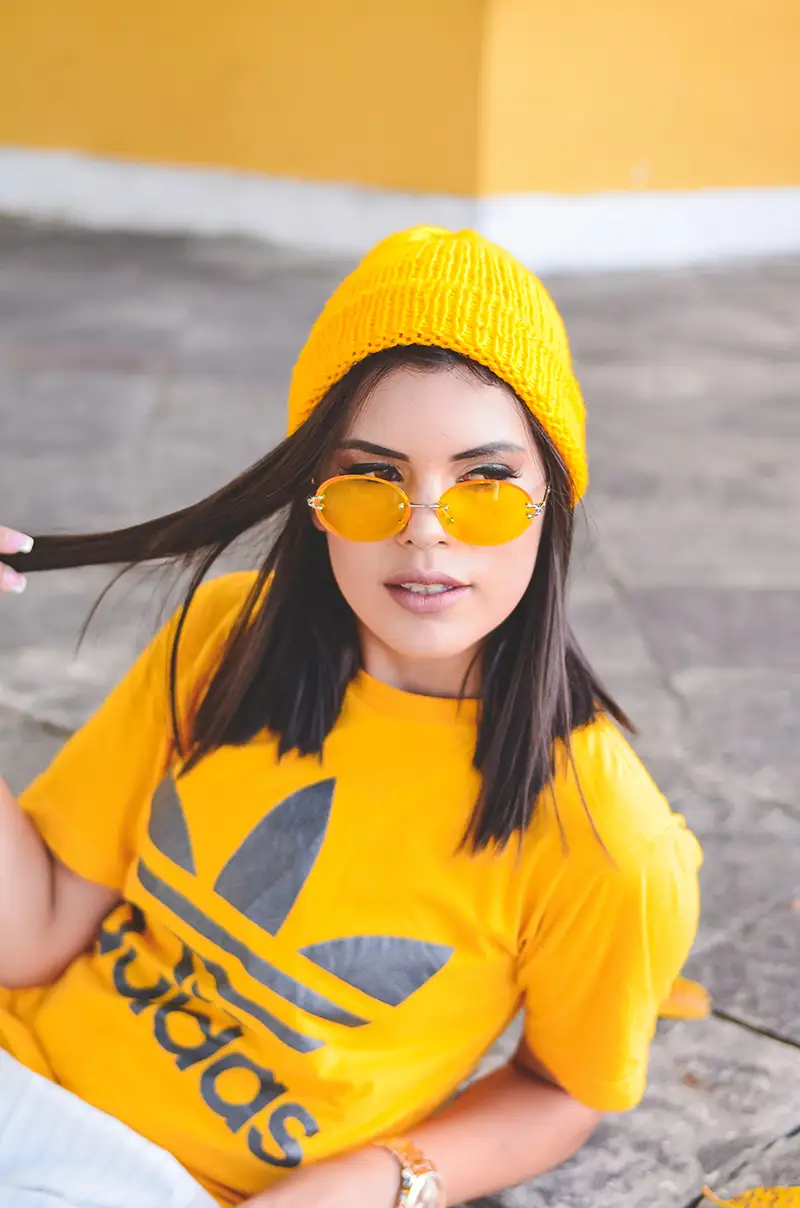 Mulher vestindo camiseta amarela com o nome Adidas na cor cinza. Usa gorro amarelo e ´coulos com lentes levemente transparentes na cor amarela.