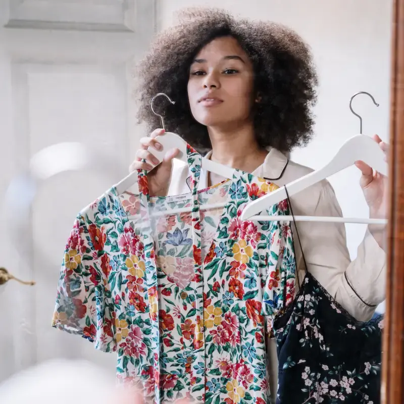 mulher negra, de cabelos cacheados, de frente a um espelho segurando duas peças de roupa para escolher o que comprar.