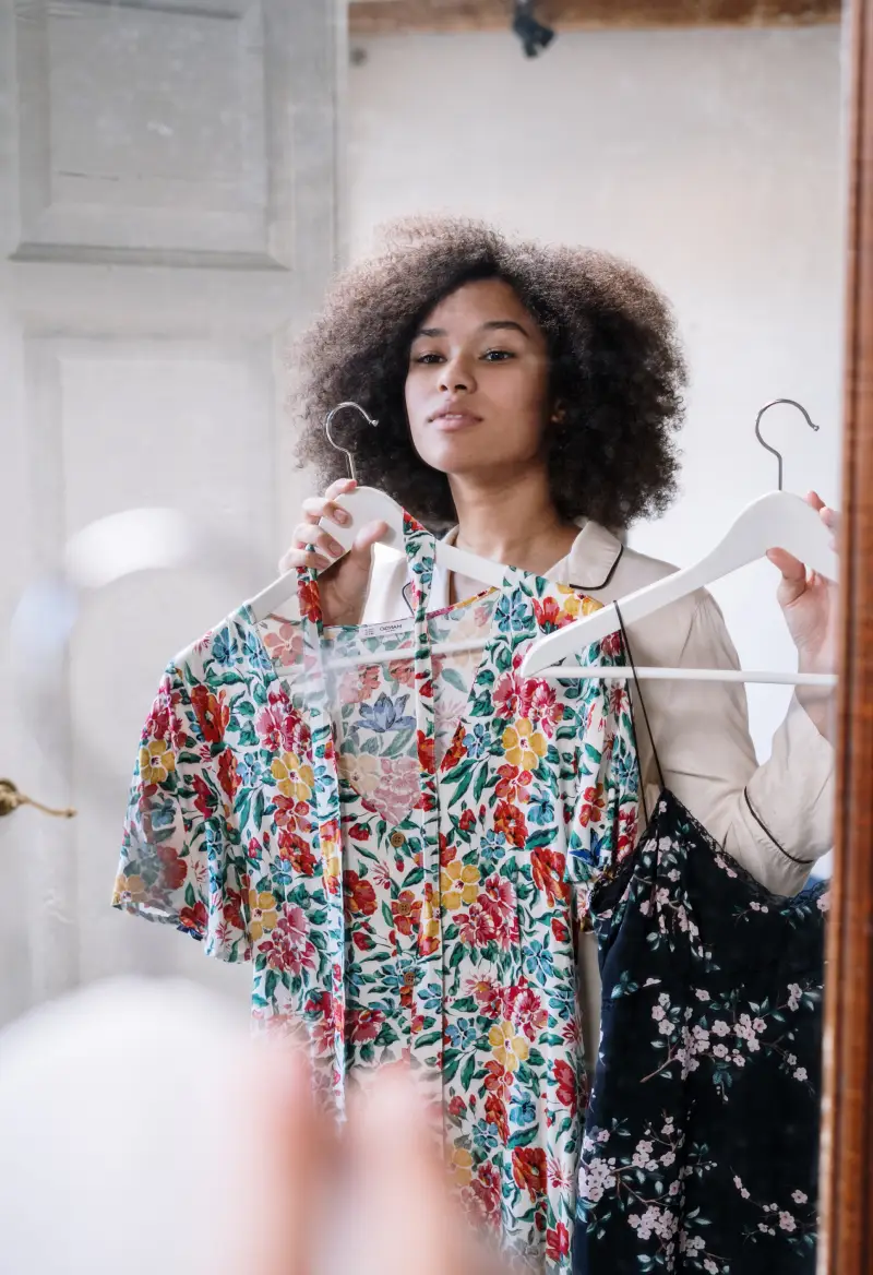 mulher negra, de cabelos cacheados, de frente a um espelho segurando duas peças de roupa para escolher o que comprar.